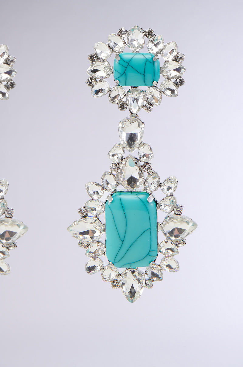 A close-up of the Gary Wood TOTALLY TURQ EARRING reveals a striking large rectangular turquoise stone at its center, framed by a detailed arrangement of clear rhinestone embellishments. Above, a smaller marbled gemstone encased in a similar design completes the elegant drop-style earring.