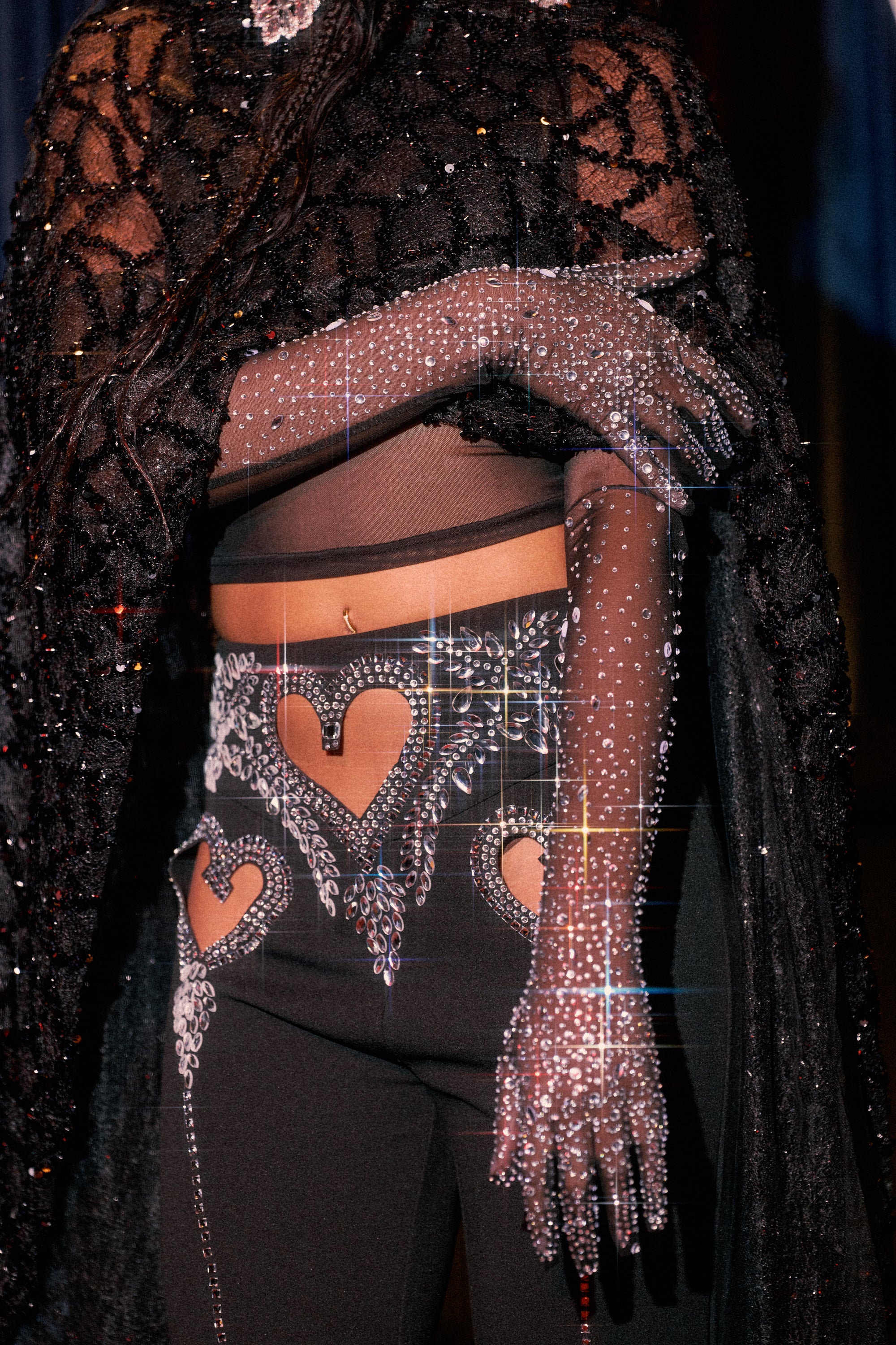 Wearing BLAIR BLING GLOVES covered in sparkling rhinestones, black pants with brown heart designs and glitter, and a dark, textured cape draped over their shoulders.