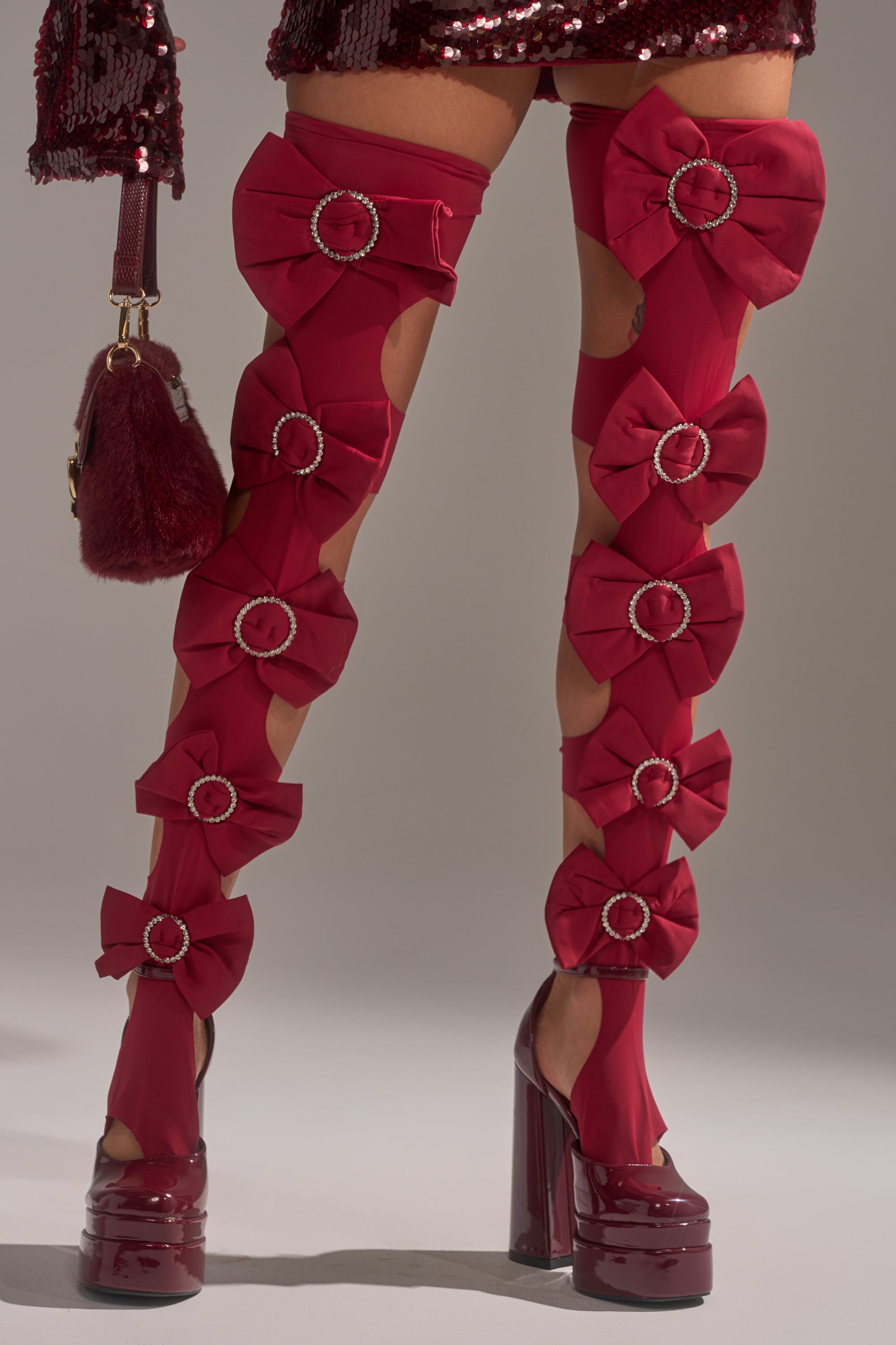 Wearing LOVES ME STIRRUP STOCKINGS—sheer red thigh-highs with large bows and rhinestone buckles—paired with glossy red platforms, a sequined skirt, and holding a burgundy fur-accented handbag. 1