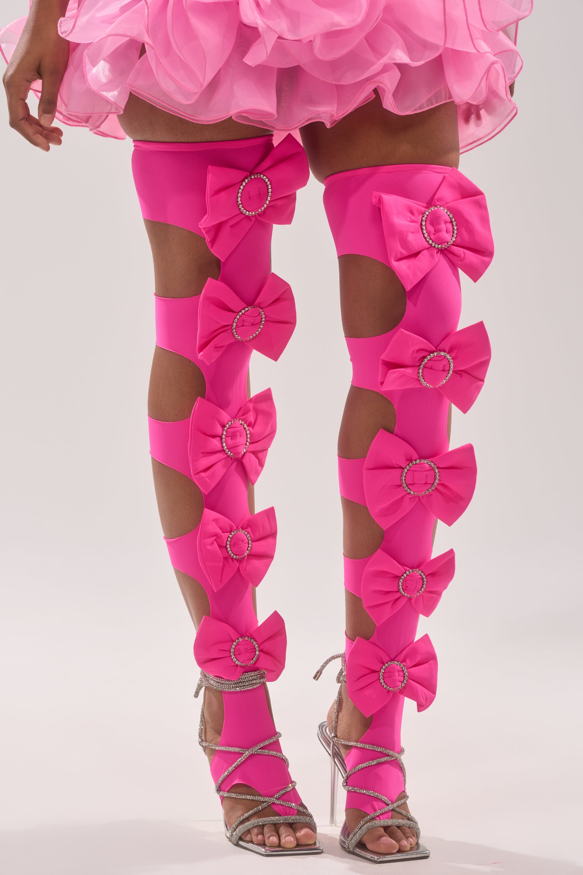 A person wears LOVE YOU TOO STIRRUP STOCKINGS in bright pink with cut-outs, large bows, and rhinestones, paired with silver strappy high-heeled sandals and a pink ruffled skirt. 2