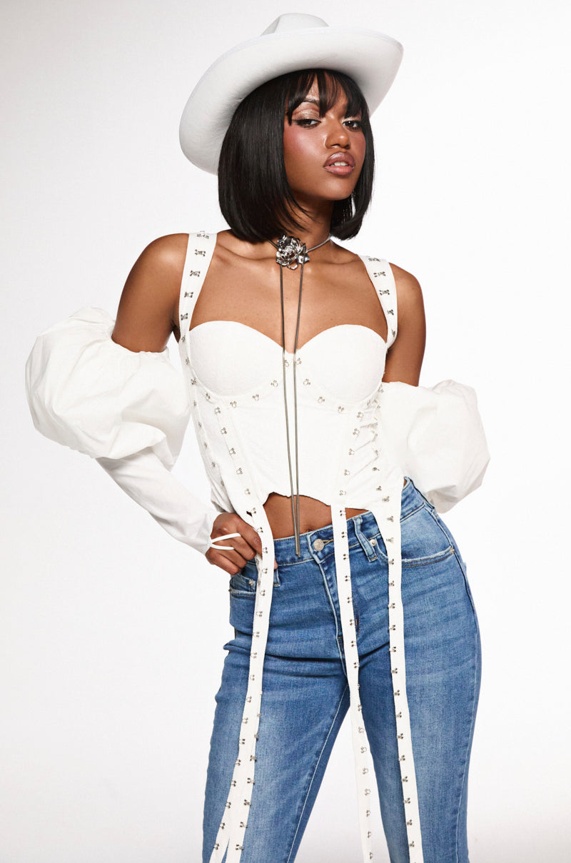 A person wears a cowboy hat, the "DONT THINK ABOUT IT LACE CORSET" with structural boning and studded details, long white sleeves, blue jeans, suspenders, and a bolo tie. They stand hand on hip against a plain white background.