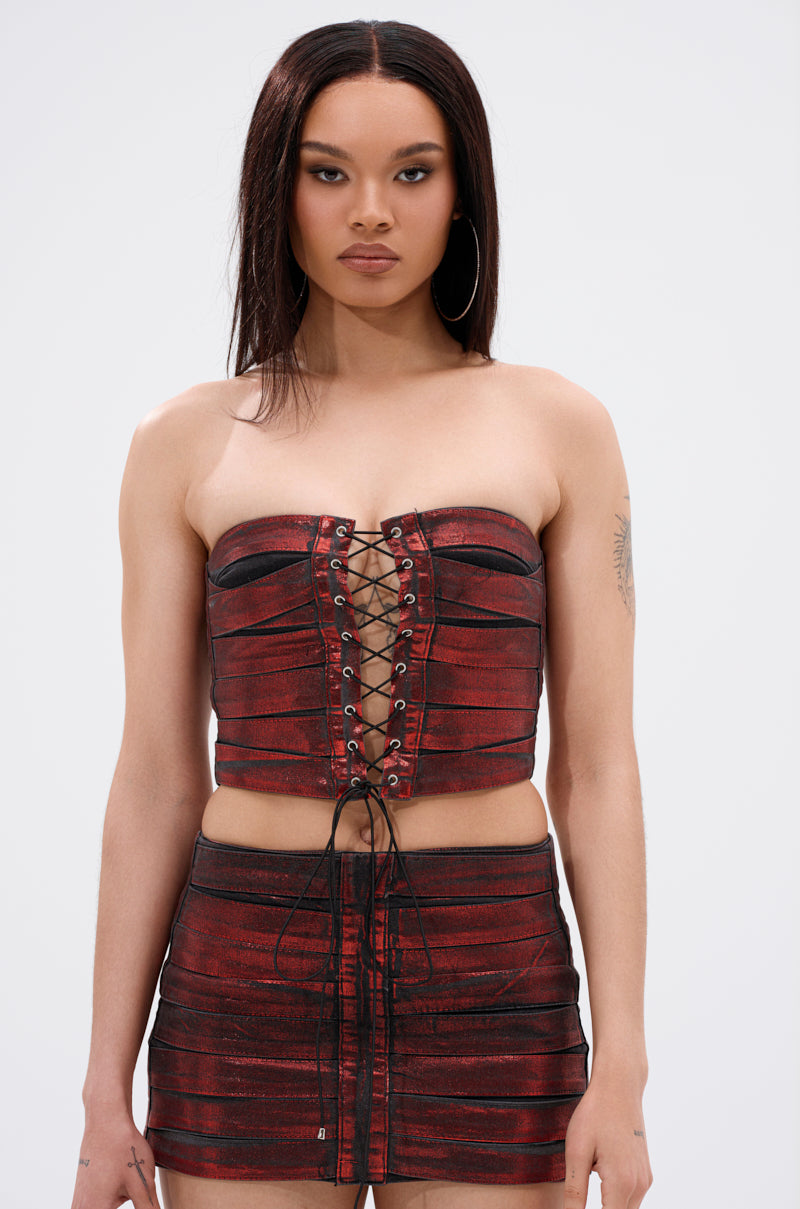 A woman with straight dark hair wears the ELECTRIC METALLIC CORSET, a strapless crop top with a lace-up front, paired with a matching mini skirt. Against a plain light background, her hoop earrings and tattoo accentuate the bold, edgy look. 1