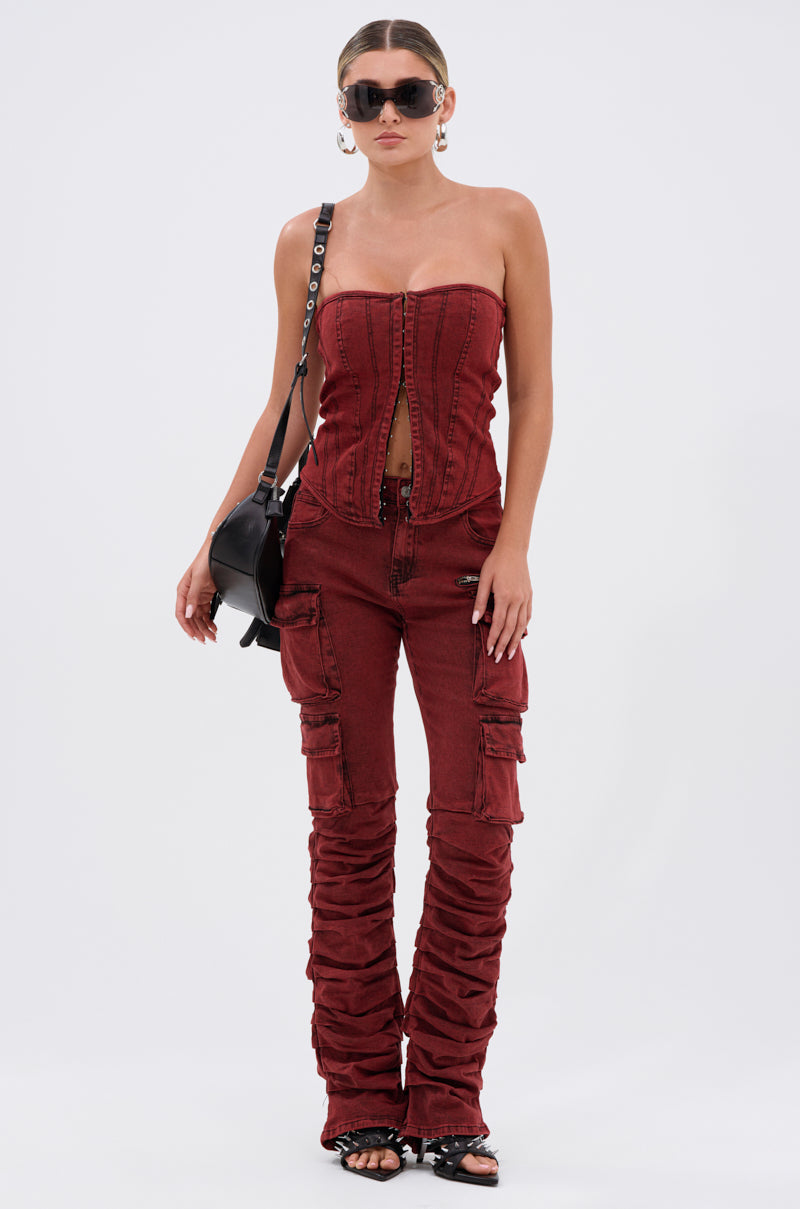 A woman stands against a plain background wearing the IT GETS EASIER STACKED CARGO PANT IN BURGUNDY with a matching strapless red denim top, sunglasses, hoop earrings, a black shoulder bag, and black heeled sandals.