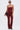 A woman stands against a plain background wearing the IT GETS EASIER STACKED CARGO PANT IN BURGUNDY with a matching strapless red denim top, sunglasses, hoop earrings, a black shoulder bag, and black heeled sandals.