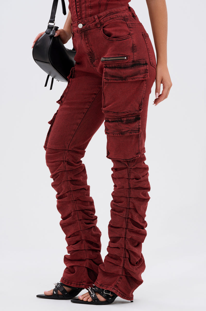 A person wears the IT GETS EASIER STACKED CARGO PANT IN BURGUNDY with ruched legs, black zipper accents, and black heeled sandals, holding a small black handbag against a plain light gray background for a streetwear vibe.