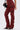 A person wears the IT GETS EASIER STACKED CARGO PANT IN BURGUNDY with ruched legs, black zipper accents, and black heeled sandals, holding a small black handbag against a plain light gray background for a streetwear vibe.