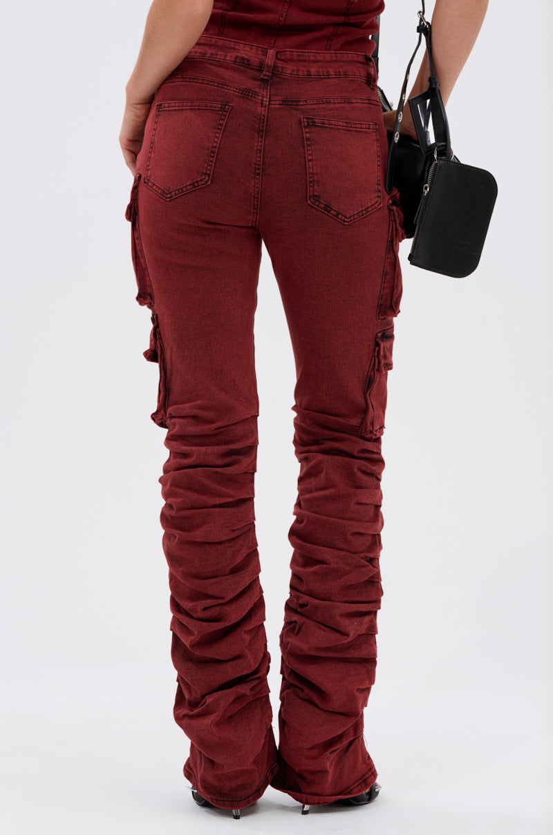 Facing away, a person models the IT GETS EASIER STACKED CARGO PANT IN BURGUNDY—high-rise streetwear denim with back pockets, stacked cargo details, and flared bottoms—paired with a small black purse for a bold statement.