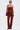 A woman in sunglasses, a strapless red denim corset top, IT GETS EASIER STACKED CARGO PANT IN BURGUNDY, black sandals, hoop earrings, and a black shoulder bag stands against a plain white background.