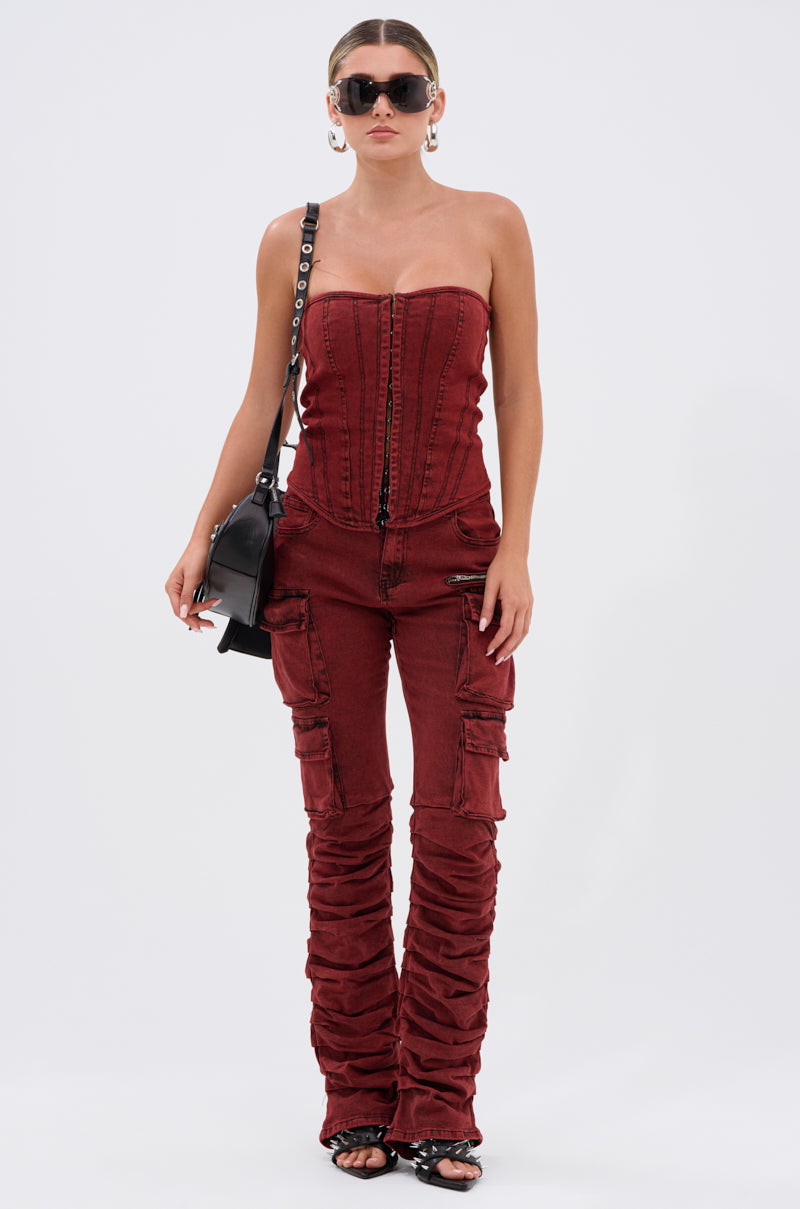 A woman wears the IT GETS EASIER STACKED CARGO PANT IN BURGUNDY, styled with black strappy sandals, hoop earrings, sunglasses, and a black shoulder bag, standing against a plain white background.
