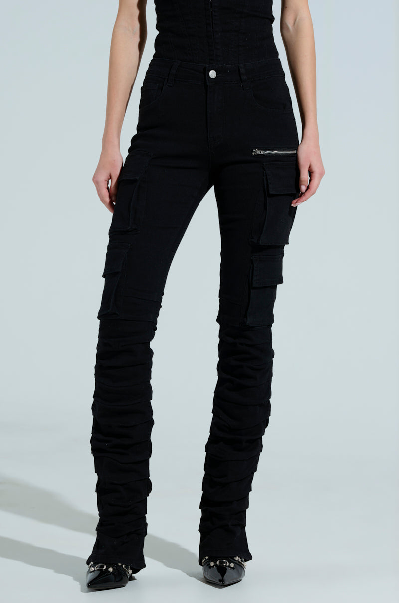 A person models black high-rise pants from the Double Vision Vintage Wash Denim Pant collection by AKIRA, featuring multiple pockets, a ruched leg design, and zippered waist pockets. The outfit is completed with black pointed-toe shoes against a light backdrop.
