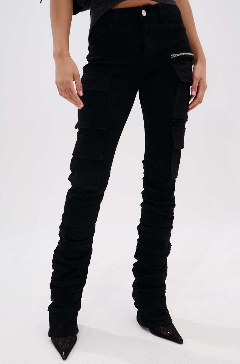 A person wears DOUBLE VISION VINTAGE WASH DENIM PANT, featuring a high-rise fit, multiple cargo pockets, and leg ruching, paired with pointed black shoes, standing against a plain white background.