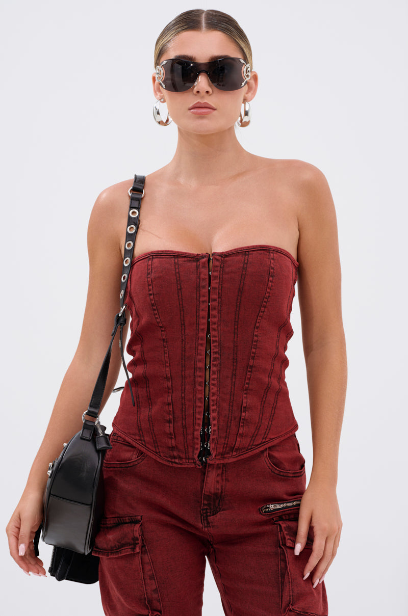 A woman faces forward wearing the IT GETS EASIER DENIM CORSET IN BURGUNDY with matching red denim pants, large sunglasses, a black shoulder bag, and large hoop earrings.