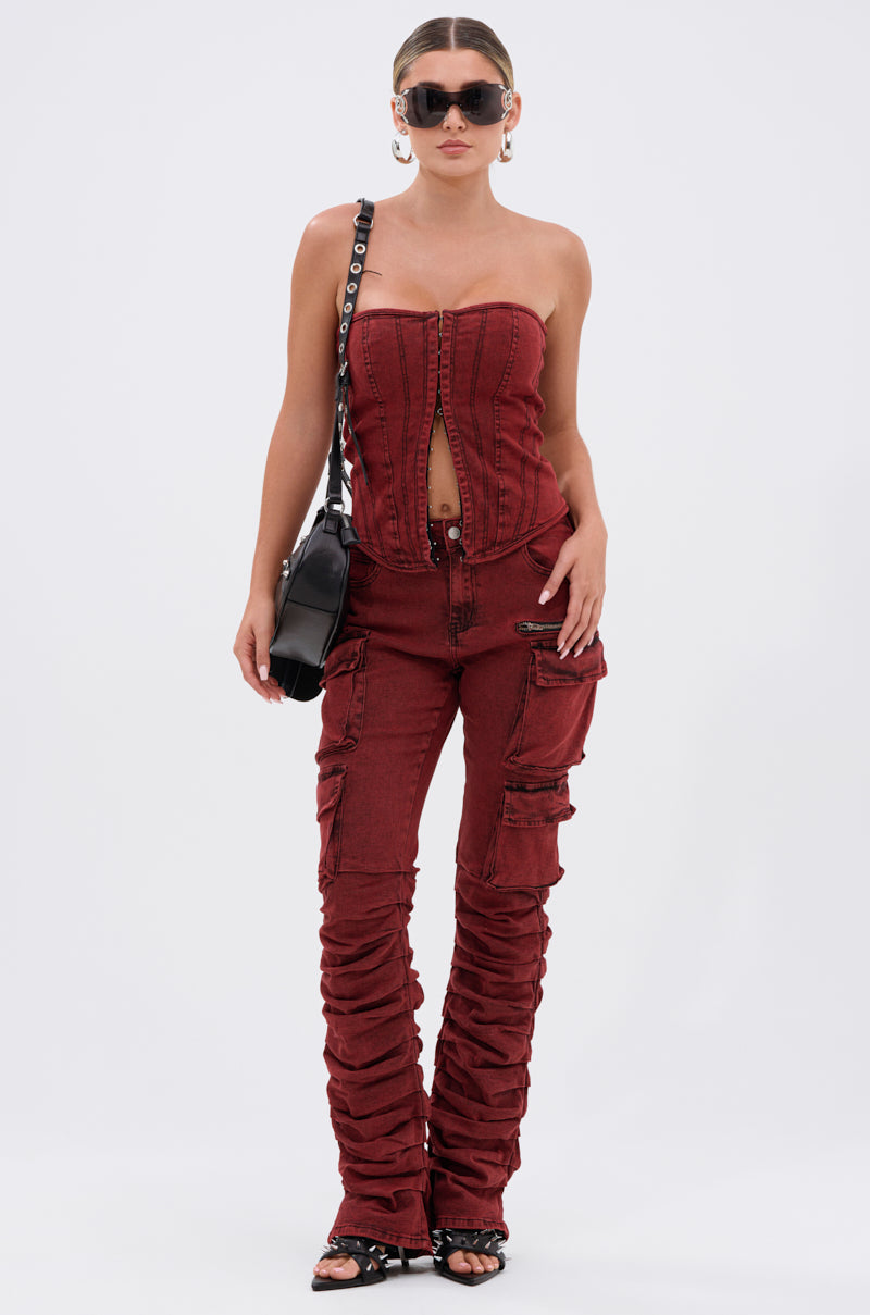 A woman wearing large sunglasses, hoop earrings, the IT GETS EASIER DENIM CORSET IN BURGUNDY, matching ruched cargo pants, black sandals, and a black shoulder bag stands against a plain white background.