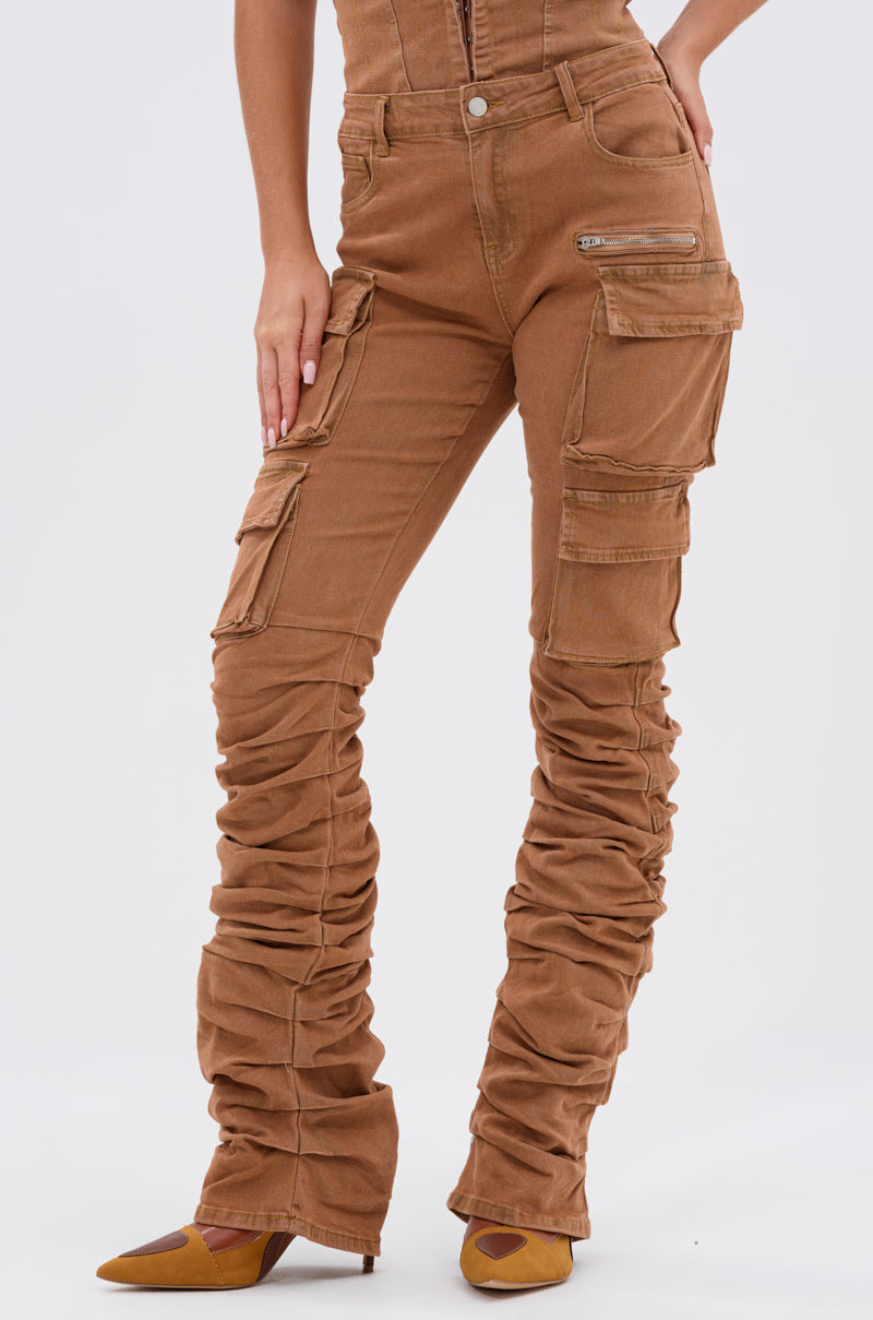 Someone wears the IT GETS EASIER STACKED CARGO PANT IN MOCHA with multiple pockets, zipper pocket, fitted waist, and ruched details, paired with pointed-toe brown shoes. Their hand is resting on their hip. 1