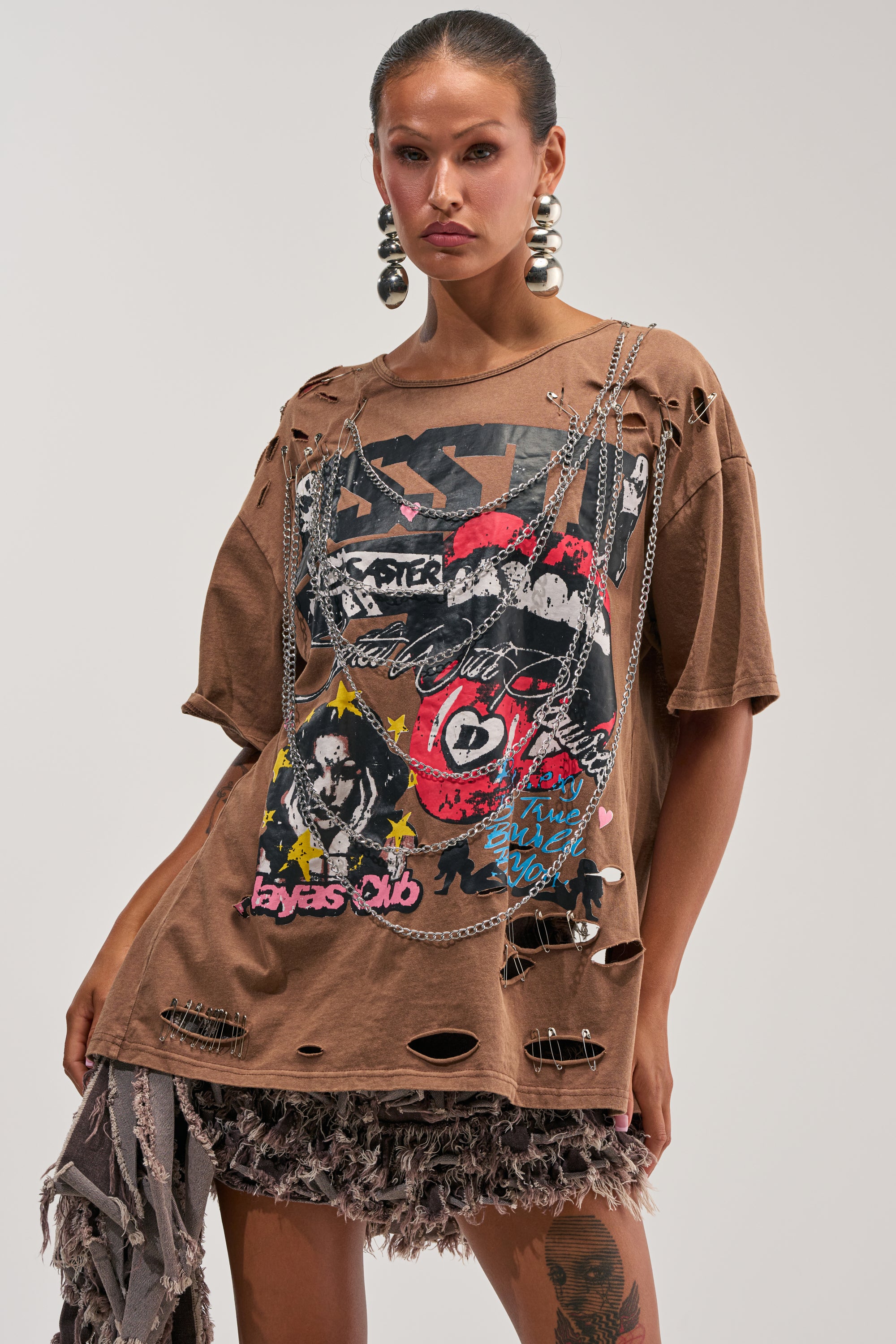A woman stands confidently against a plain background, wearing the CHAOS CLUB OPEN BACK GRAPHIC TEE—an oversized, distressed graphic top—paired with layered necklaces, large earrings, and a textured skirt.