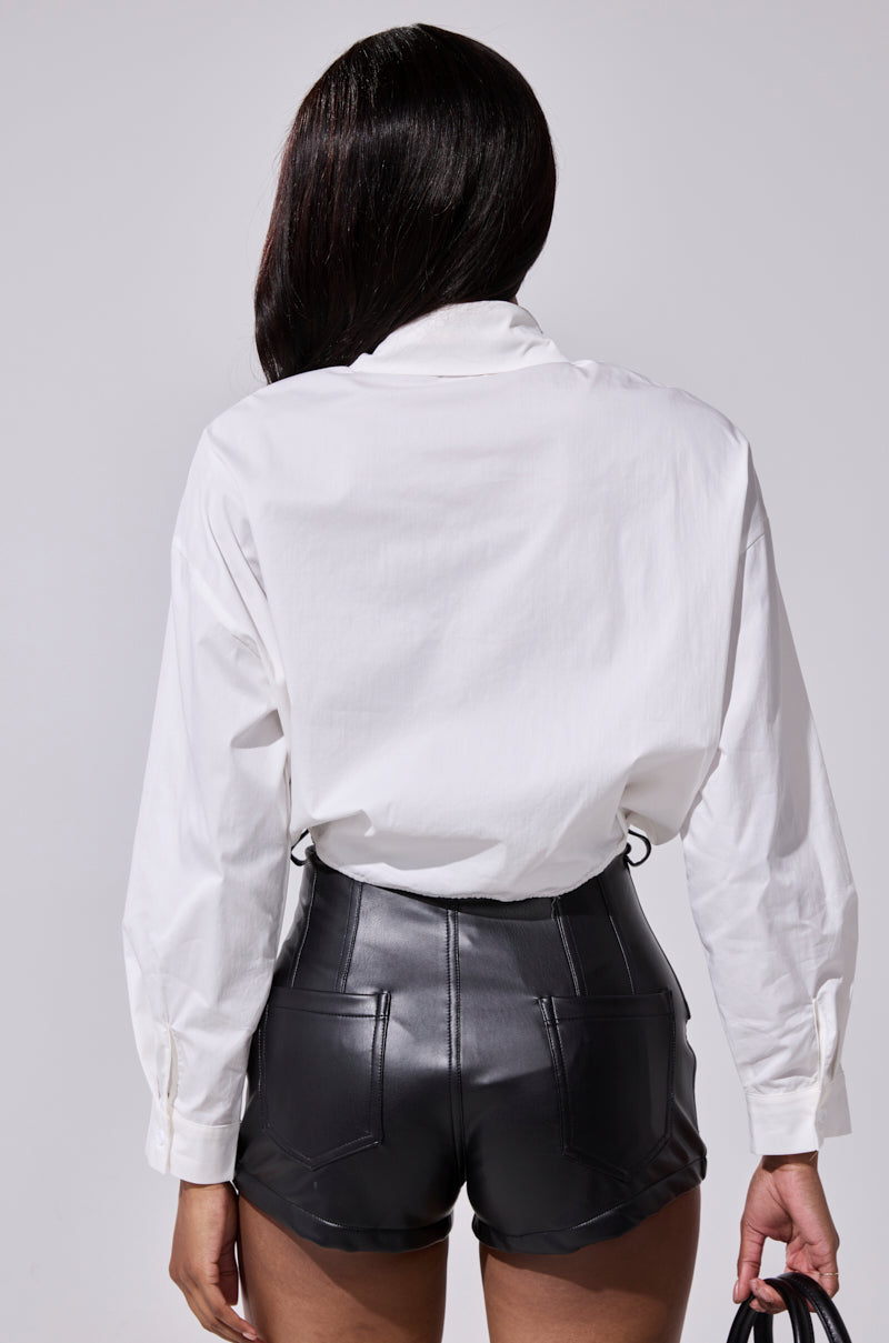 A person with long dark hair faces away, wearing the LETS TAKE IT OFFLINE BUTTON DOWN shirt tucked into high-waisted black faux leather shorts, holding a small black item in their left hand against a plain, light background. 4