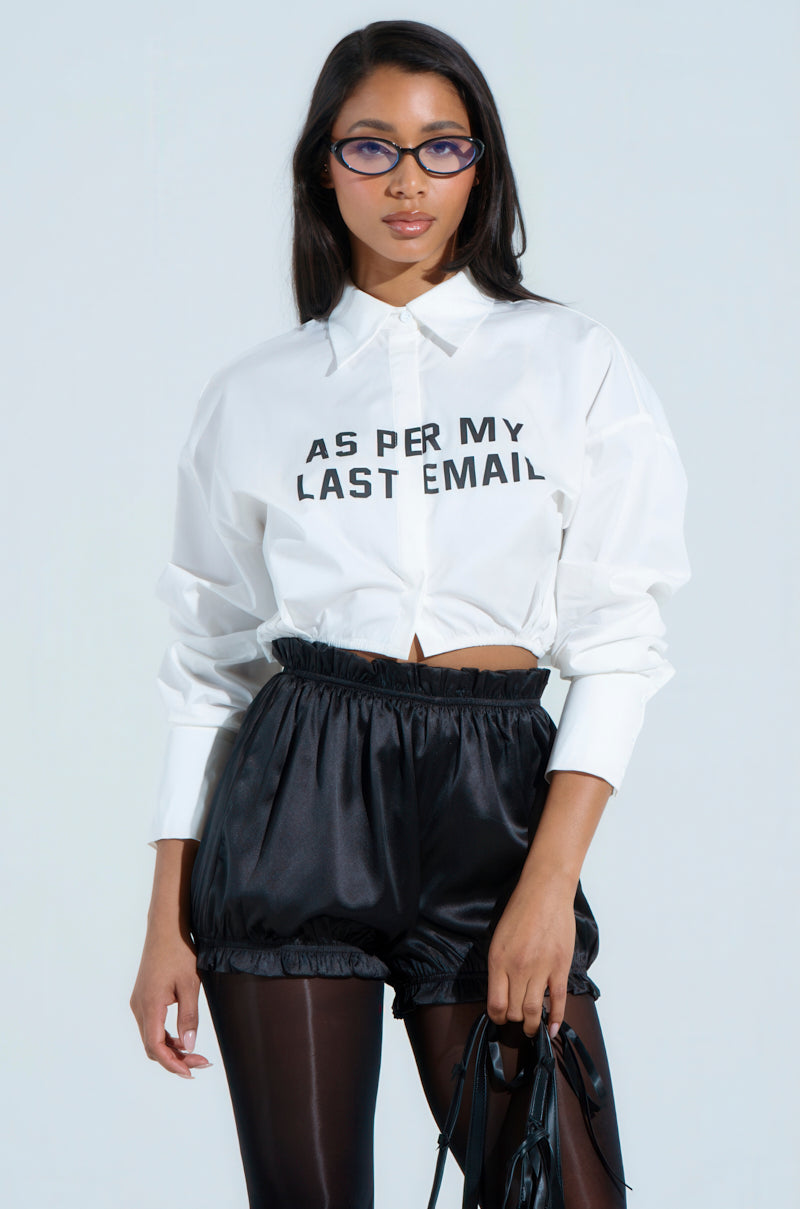 A woman wearing blue-tinted glasses and a BUSINESS CASUAL BUTTON UP inscribed with "AS PER MY LAST EMAIL" pairs her statement top with high-waisted black shorts and sleek black tights, embodying a chic business casual look against a plain background.