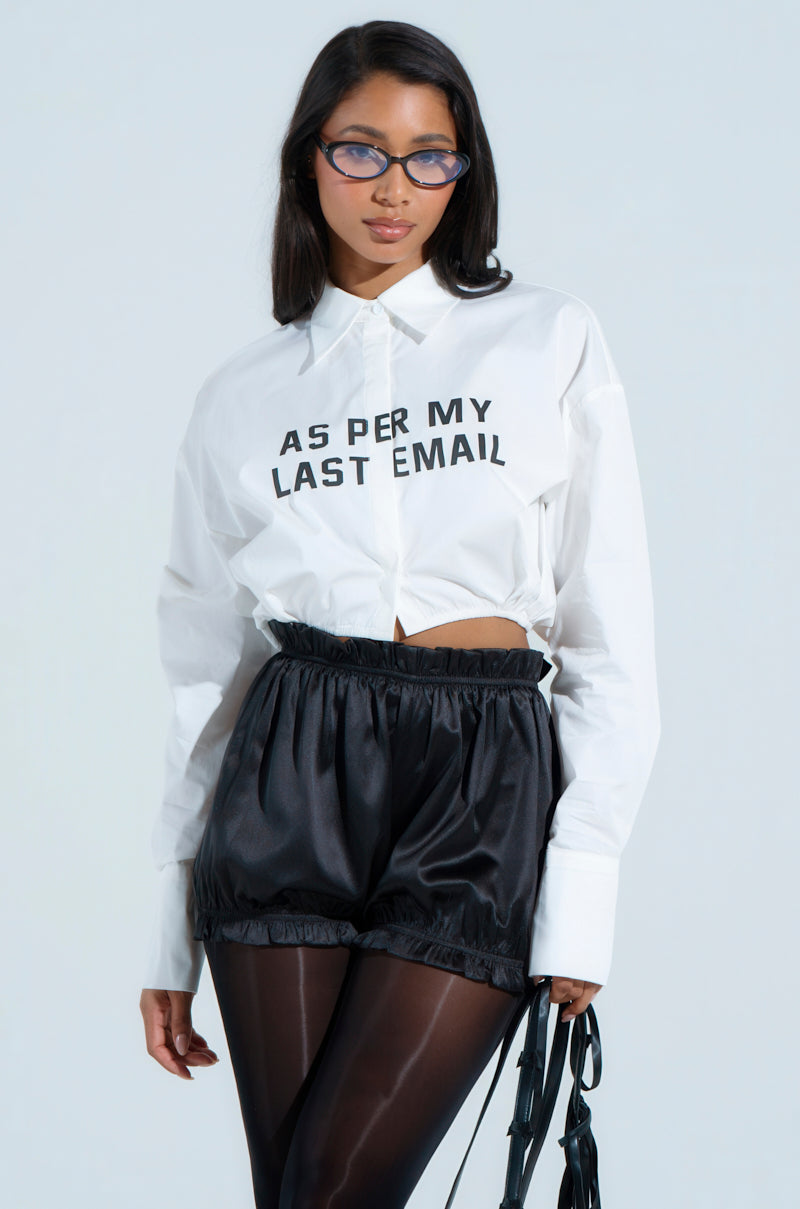 A woman with long hair wears BUSINESS CASUAL BUTTON UP cat-eye glasses, and a white cotton-blend blouse saying "AS PER MY LAST EMAIL." She pairs it with black satin shorts and sheer black tights, creating a business casual look against a plain light grey background.