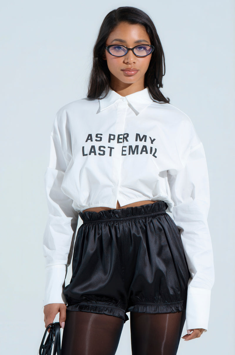 A person with long hair and glasses wears a BUSINESS CASUAL BUTTON UP blouse featuring "AS PER MY LAST EMAIL." They style it with black business casual shorts over tights and hold a sleek black bag, set against a plain backdrop to highlight their outfit.