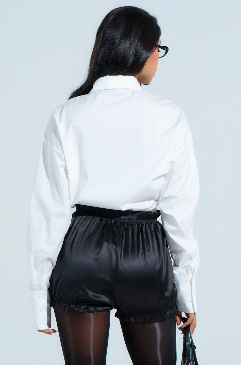 A person with long dark hair faces away, wearing the BUSINESS CASUAL BUTTON UP—a white cotton-blend blouse—paired with black high-waisted satin shorts and dark tights for a chic business casual look. They hold a small bag and wear glasses against a plain light backdrop.