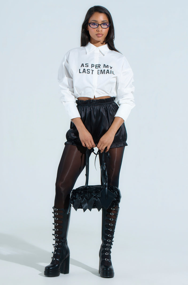In a crisp business casual look, a person confidently wears the "BUSINESS CASUAL BUTTON UP," featuring "AS PER MY LAST EMAIL" text, paired with black high-waisted shorts, sheer black tights, knee-high lace-up boots, glasses, and a black handbag against a light blue background.