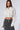 A woman with long brown hair is wearing a white MINIMAL ELEGANCE Button Down Poplin Shirt with extra long sleeves and a drawstring waist, paired with a high-waisted gray skirt. She stands against a plain white background, exuding minimal elegance as she looks directly at the camera.