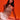A woman with long dark hair poses against an orange background, wearing the ICY GIRL RHINESTONE EMBELLISHED TOP IN GOLD and white pants. She sits with one hand on the ground, gazing confidently at the camera.