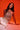 A woman with long dark hair poses against an orange background, wearing the ICY GIRL RHINESTONE EMBELLISHED TOP IN GOLD and white pants. She sits with one hand on the ground, gazing confidently at the camera.
