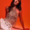 A woman with long dark hair poses against an orange background, wearing the ICY GIRL RHINESTONE EMBELLISHED TOP IN GOLD and white pants. She sits with one hand on the ground, gazing confidently at the camera.