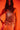 A woman exudes confidence and elegance in the ICY GIRL RHINESTONE EMBELLISHED TOP IN GOLD, styled over white satin pants against a warm orange background. Her long, wavy hair is loose, and she poses with one hand in her pocket.