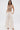 A woman stands against a white background wearing the ICY GIRL RHINESTONE EMBELLISHED TOP IN GOLD and high-waisted, wide-leg white pants. Hands in her pockets, she completes the festival-ready look with strappy gold heels.