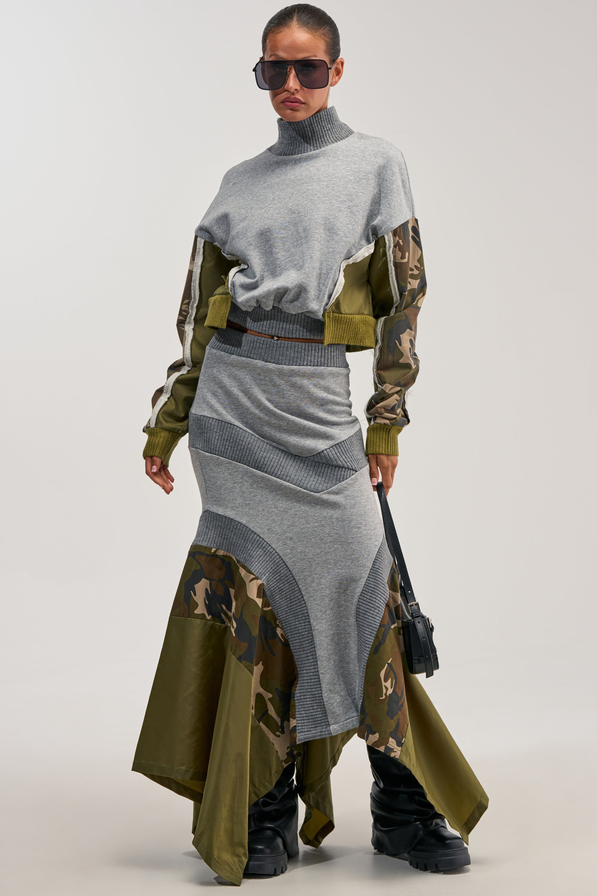 A woman stands against a plain background wearing sunglasses, a grey turtleneck sweater with camouflage accents on the sleeves, and the INES COLORBLOCKED MAXI SKIRT—a matching grey mermaid maxi skirt featuring camo details. She completes the look with chunky black boots and carries a small black bag.
