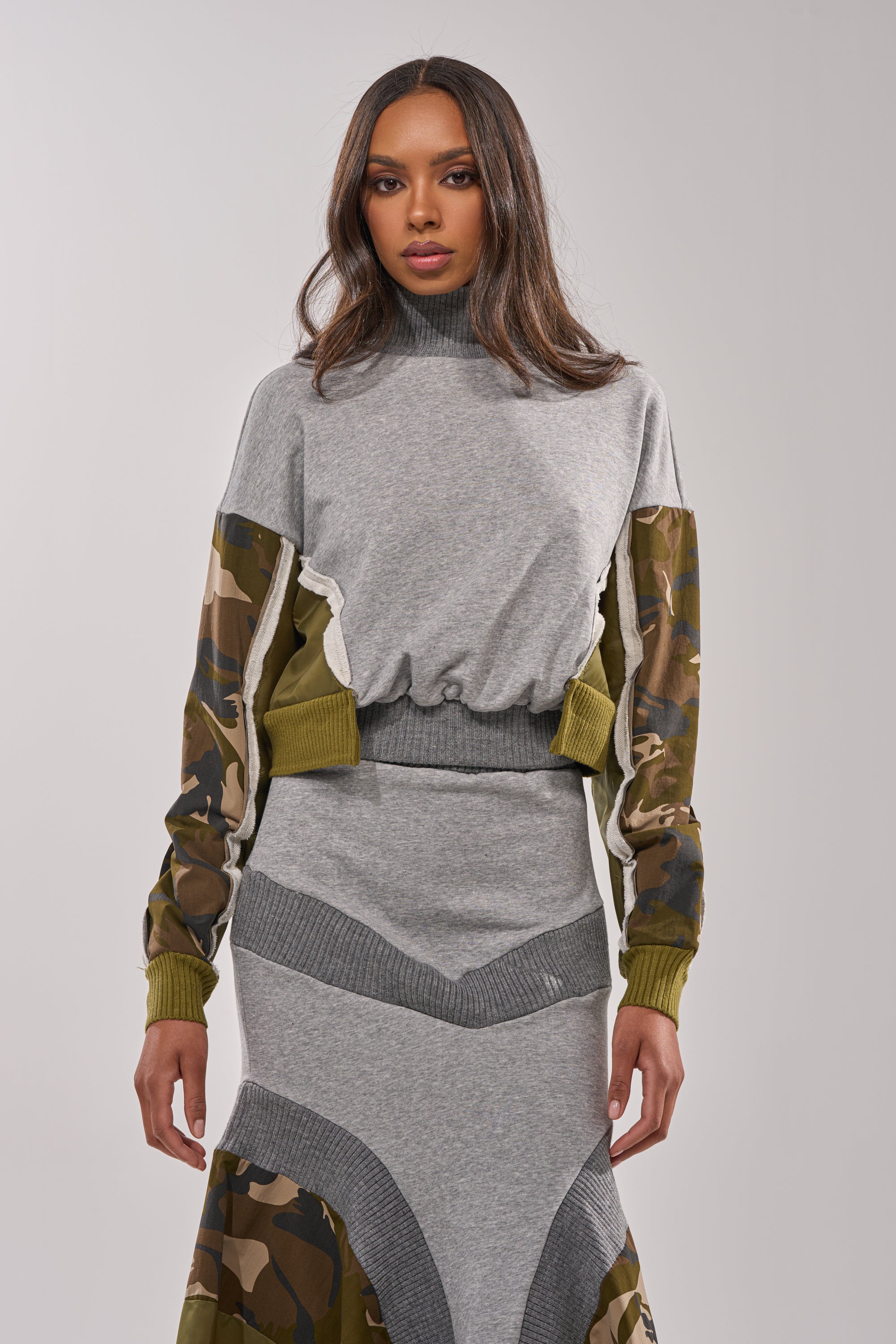 A woman stands against a plain light background, looking straight at the camera in the INES COLORBLOCKED SWEATSHIRT— a grey cropped style with camouflage-patterned sleeves—paired with a matching skirt featuring camo details. 0