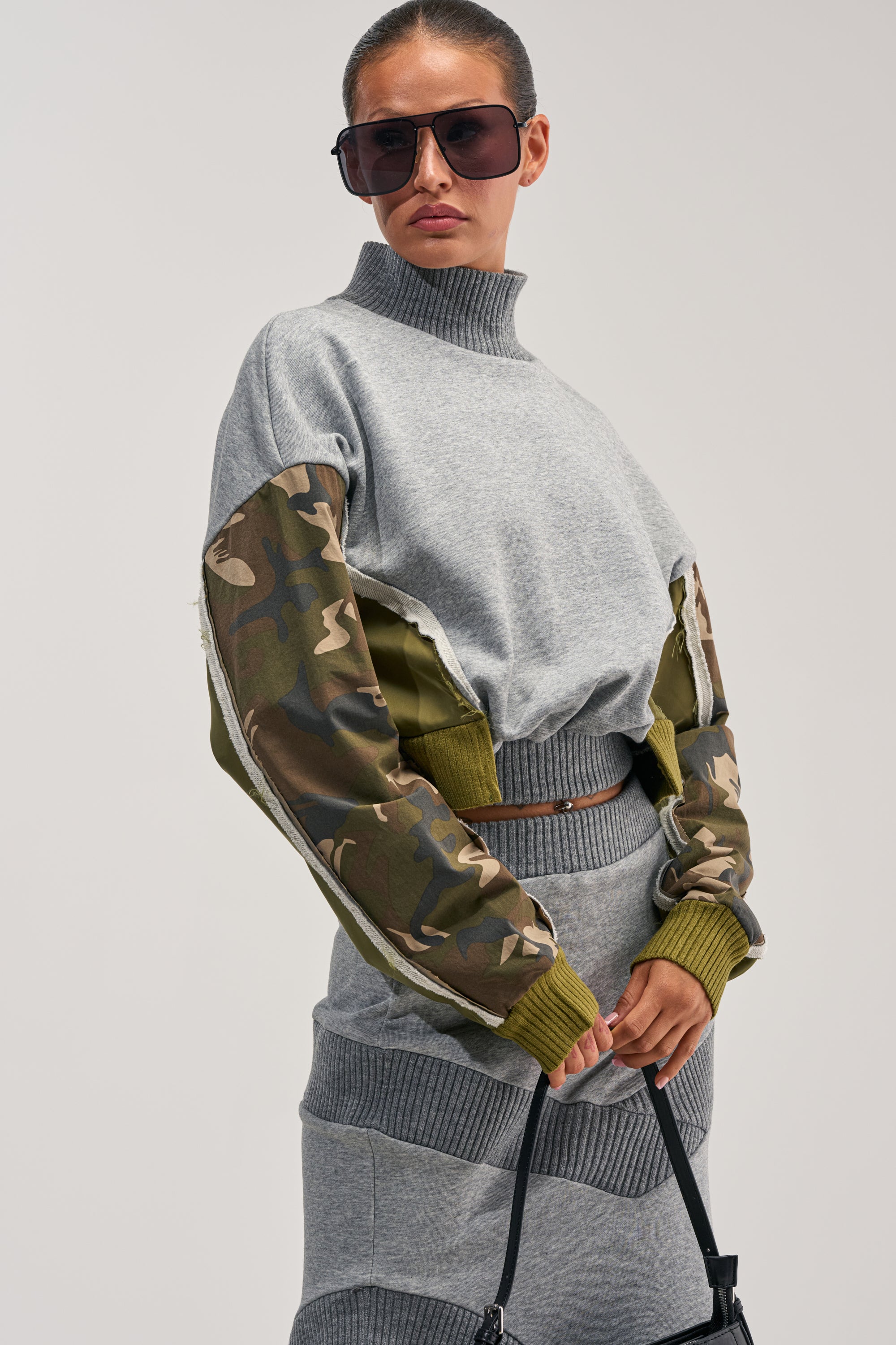 Wearing sunglasses and the INES COLORBLOCKED SWEATSHIRT—a grey turtleneck featuring camo paneling on the sleeves—paired with a matching grey skirt and holding a black handbag, she poses confidently against a light background, embodying effortless streetwear style.