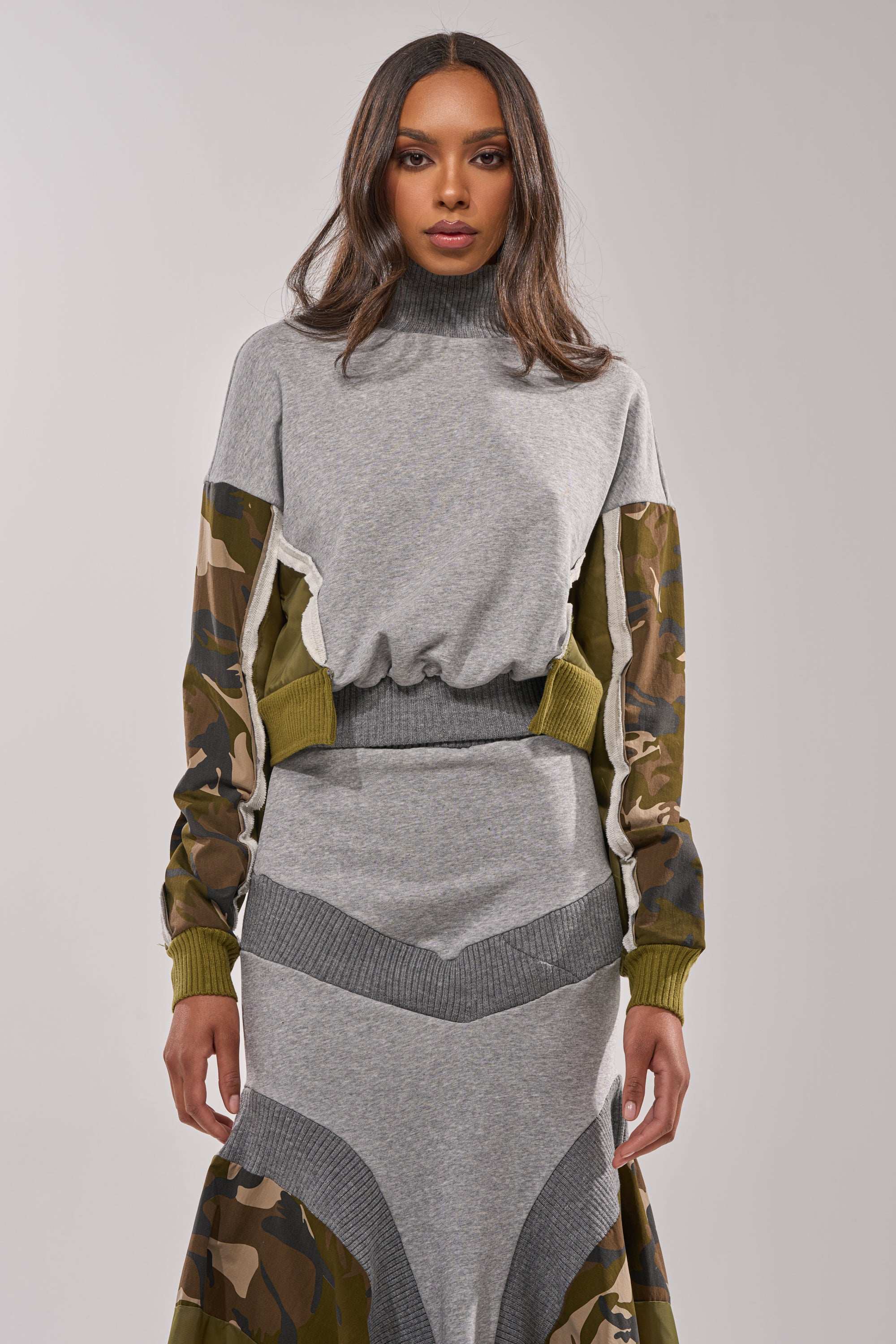 A woman faces forward in the INES COLORBLOCKED SWEATSHIRT, a grey top with ribbed cuffs and hem, camo and olive-green sleeve panels, paired with a matching skirt featuring chevron and camo patterns. 1