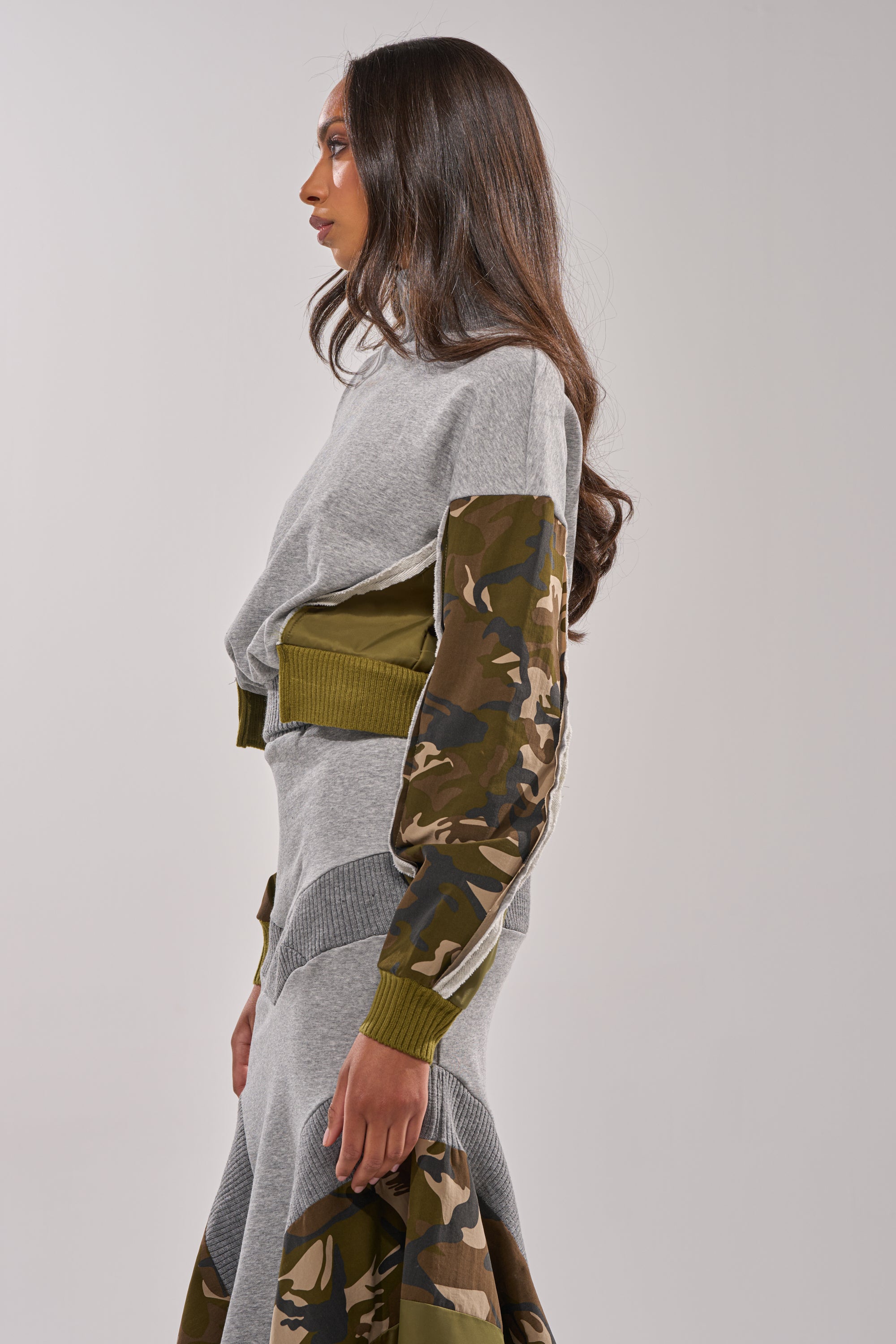 A woman stands in profile, wearing the INES COLORBLOCKED SWEATSHIRT featuring grey with green and brown camo patch sleeves. Her long dark hair drapes over her shoulders against a plain light grey background. 2