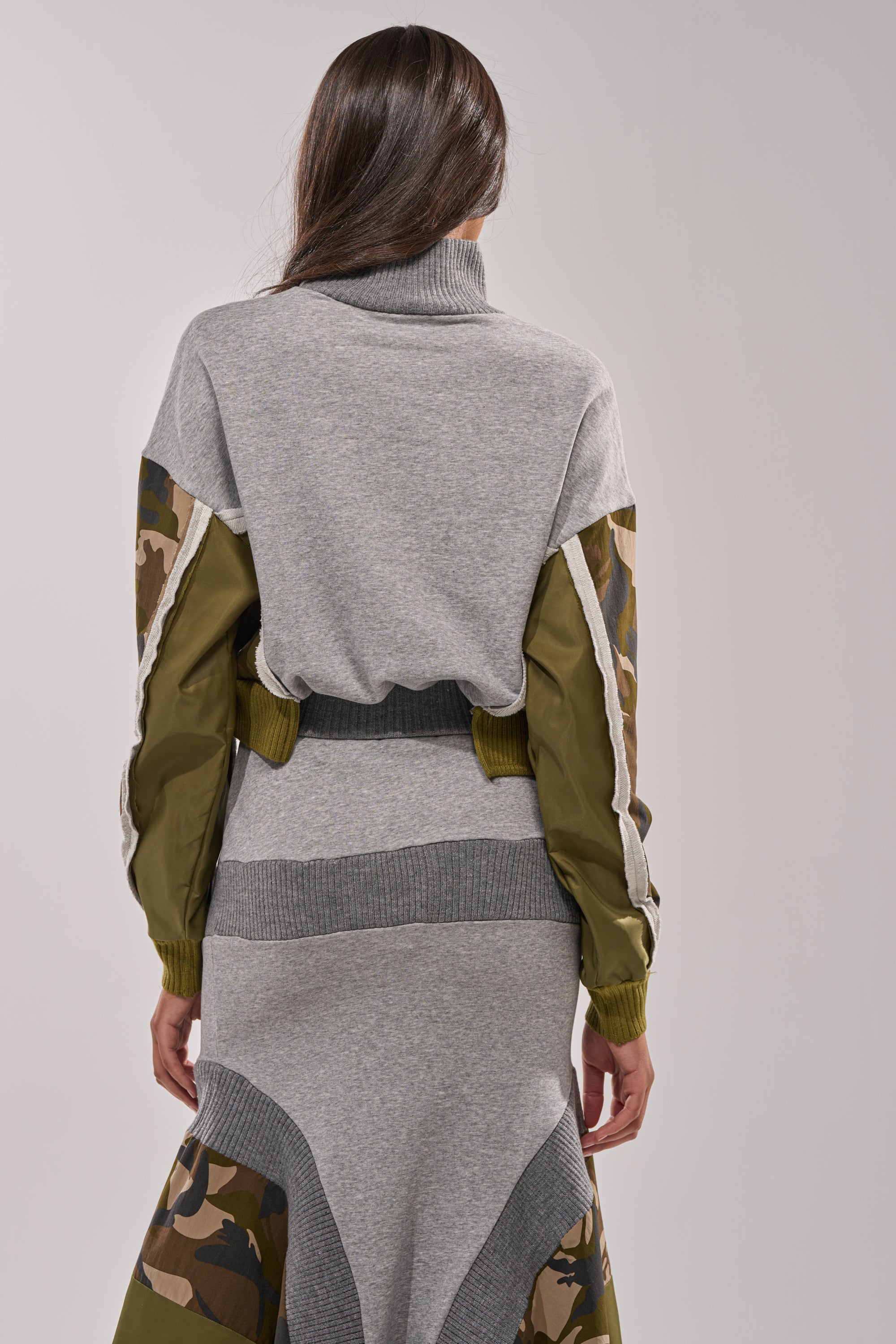 A woman with loose long brown hair wears the INES COLORBLOCKED SWEATSHIRT, featuring a gray turtleneck and olive camo sleeves, paired with a matching ribbed knit long skirt. She stands facing away. 3