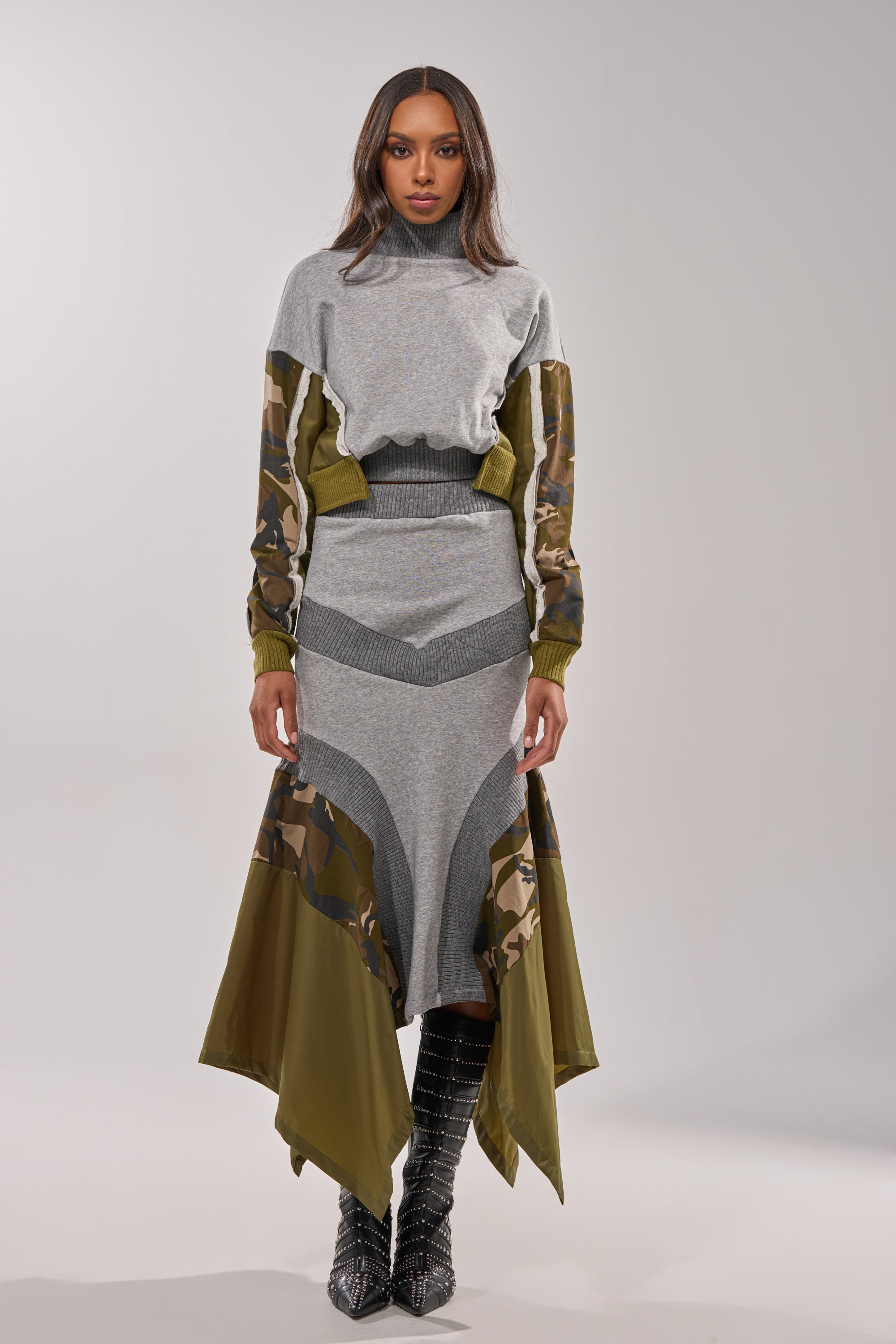A woman faces forward wearing the INES COLORBLOCKED SWEATSHIRT, a gray and olive green dress with camo-patterned sleeves, colorblocked panels, textured details, a high neckline, asymmetrical hem, and black textured boots. 4