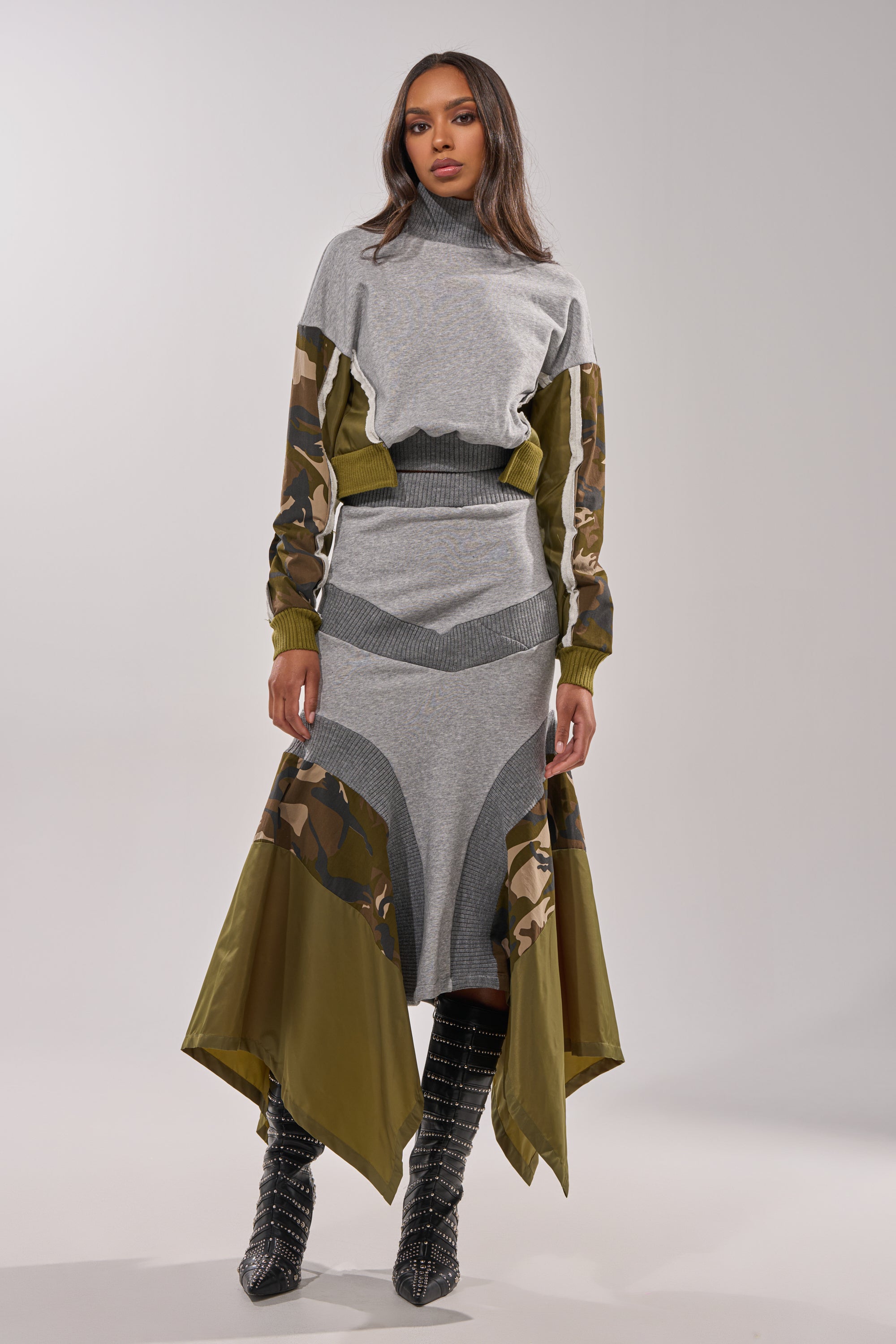 A woman models the INES COLORBLOCKED SWEATSHIRT, featuring grey colorblocking, camouflage sleeves and side panels, and an olive-green hem. She pairs it with black studded boots and poses confidently against a plain background. 5