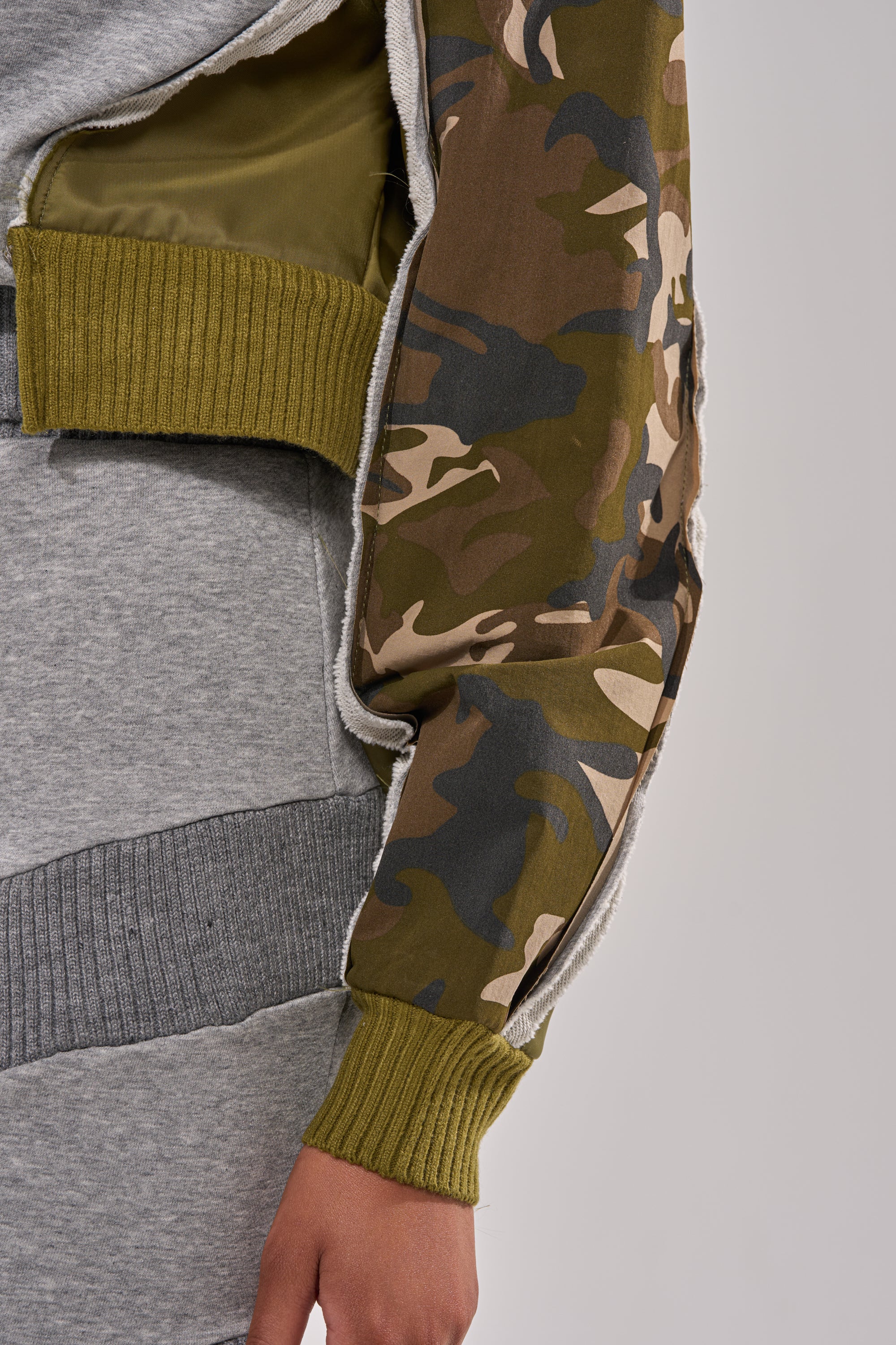 Close-up of a person wearing the INES COLORBLOCKED SWEATSHIRT, which features olive green ribbed cuffs, a camouflage-patterned sleeve, and is partially unzipped to reveal an olive green lining. Paired with a gray ribbed knit outfit. 6
