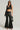 A person models a trendy ensemble showcasing the YES PLEASE FAUX LEATHER PANT, characterized by its high-rise fit and flared pantlegs. They pair it with a chic black, single-sleeve crop top and accessorize with sunglasses, gloves, a small handbag, and a decorative chain belt.