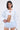 A woman with straight dark hair wears the TALK TO ME NICE CORSETED JACKET IN WHITE over a white crop top and light blue lace ruffled shorts. She stands against a plain light background, looking confidently at the camera.