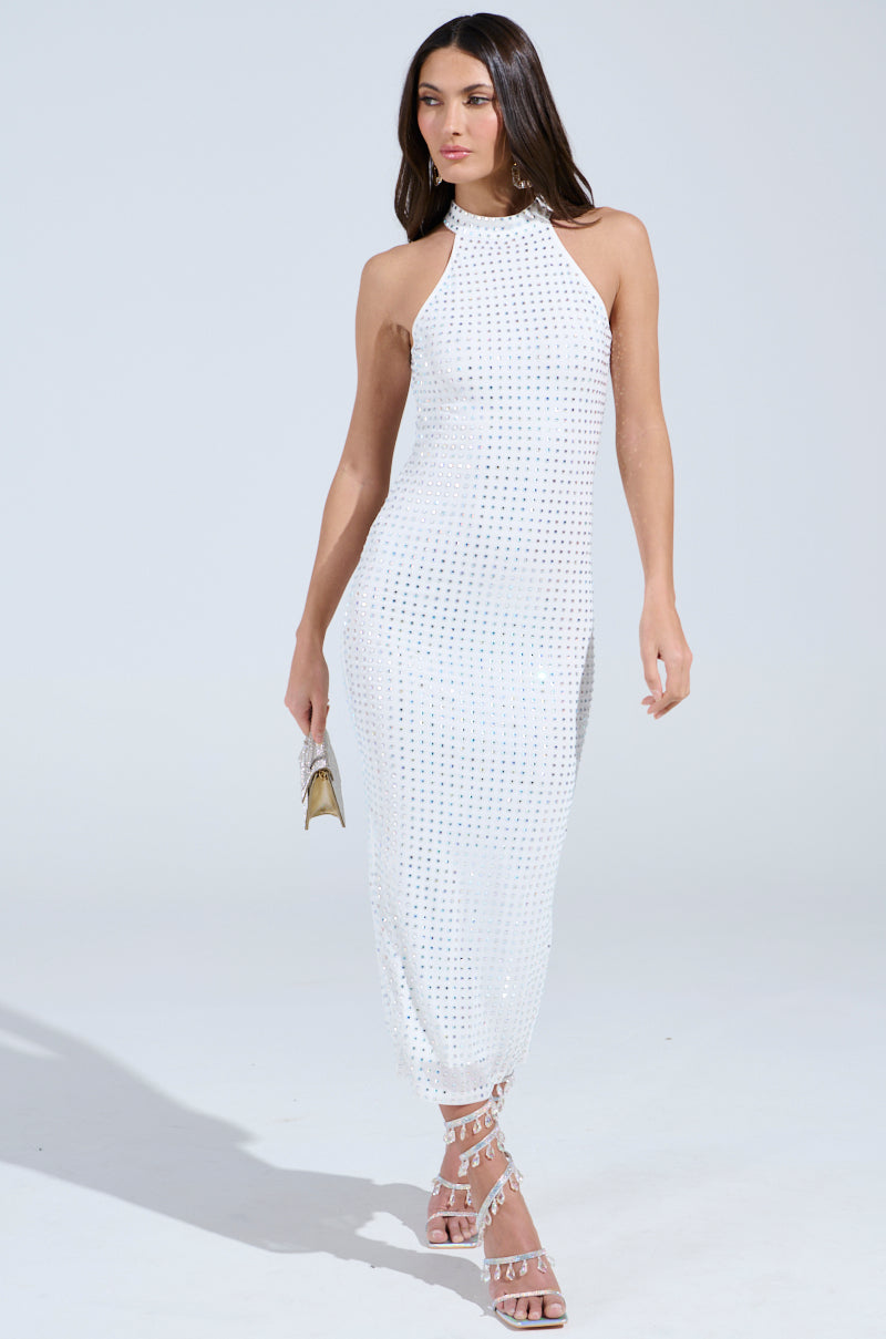 Elegantly dressed in the sleeveless, halter-neck TAKE ME OUT RHINESTONE MIDI DRESS with a textured pattern and open back, a woman carries a small gold clutch. She walks in strappy white high-heeled sandals against a plain, light-colored background.