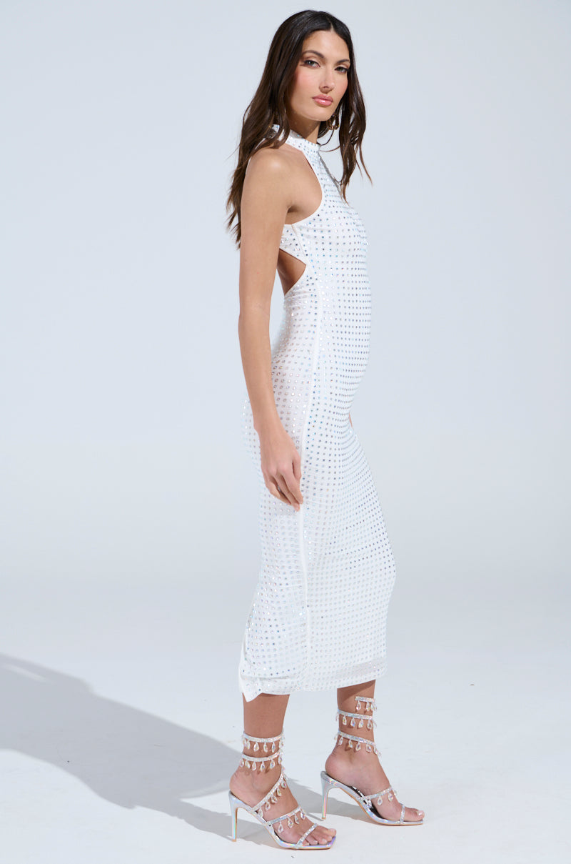 A woman models the TAKE ME OUT RHINESTONE MIDI DRESS, a sleeveless, form-fitting white dress featuring cut-out sides, an open-back design, and a grid of small metallic studs. She wears strappy high-heeled sandals against a plain light grey background.