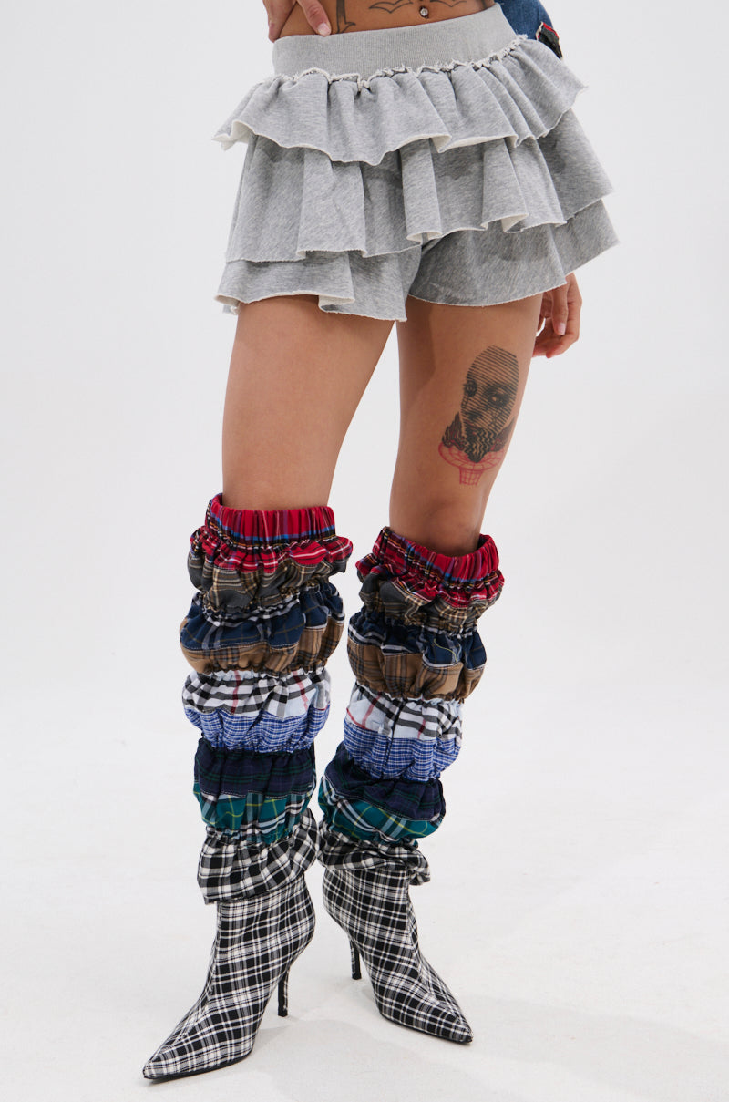 A person wearing a layered grey ON THE MOVE RUFFLE SKORT, colorful plaid leg warmers, black-and-white plaid high-heeled boots, and showcasing a thigh tattoo stands against a plain white background.