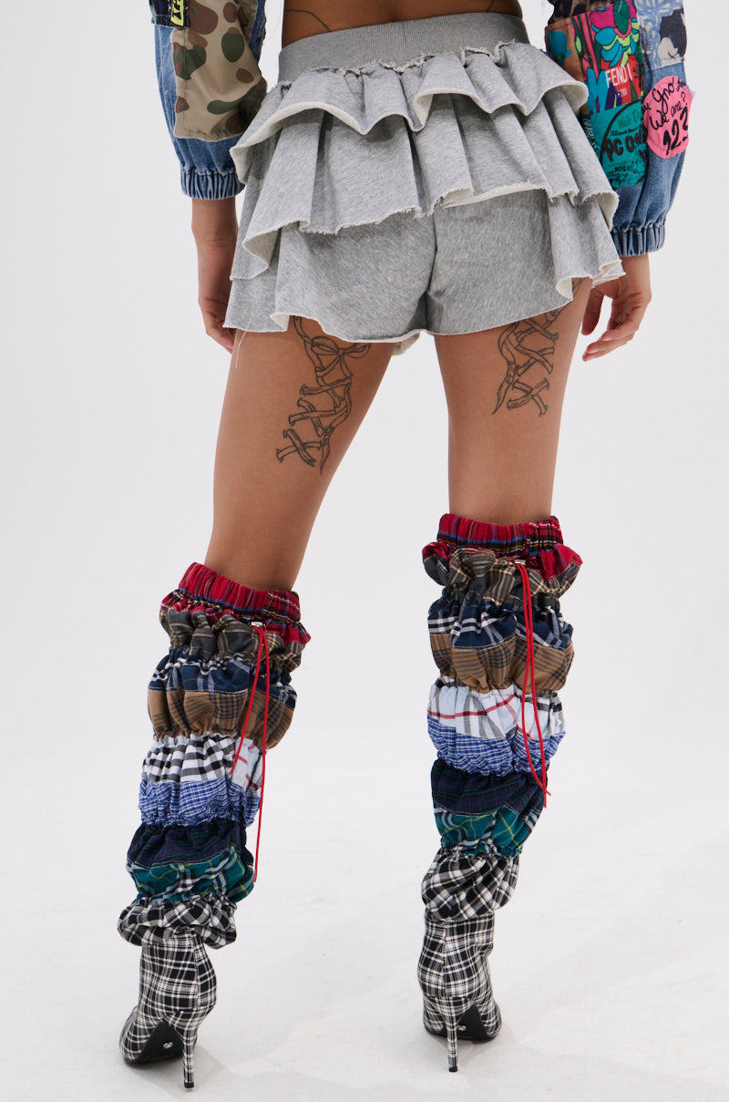 A person stands facing away, wearing the ON THE MOVE RUFFLE SKORT, vibrant printed sleeves, and tall patchwork leg warmers over plaid high-heeled boots. Tattoos are visible on the backs of their thighs.