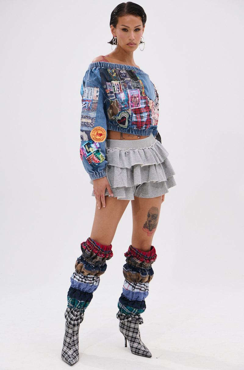 A woman poses in a colorful denim patchwork top, ON THE MOVE RUFFLE SKORT in gray, plaid mixed-pattern leg warmers, and plaid pointed-toe boots against a plain white background.