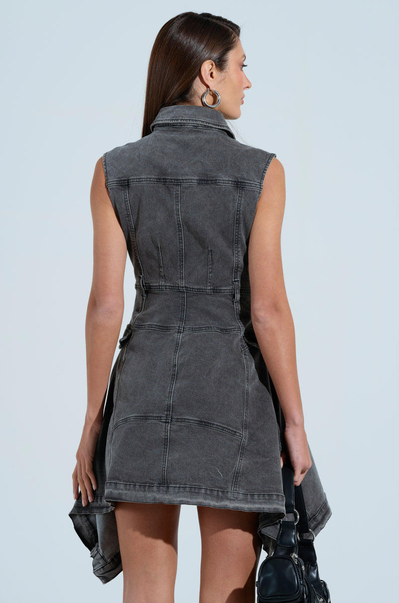 A woman with long brown hair is seen from behind, wearing a MICHELLE DENIM MINI DRESS in dark gray. The sleeveless dress has a fitted waist, flared hem, and button front closure. She carries a black bag in her right hand against the light blue background.