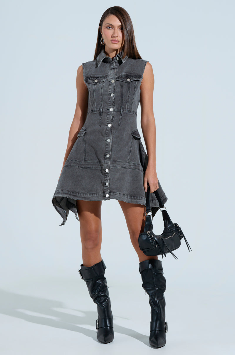 Wearing the MICHELLE DENIM MINI DRESS, a person stands confidently in a sleeveless design with button front closure. They pair it with black knee-high boots and hold a small black bag against a plain light blue background, accentuating the stylish outfit.