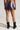 A person is facing away from the camera, dressed in the SILENCE MINI SKIRT with an abstract wavy design, paired with black boots. The plain, light-colored backdrop accentuates the modern low-rise shape of their outfit.